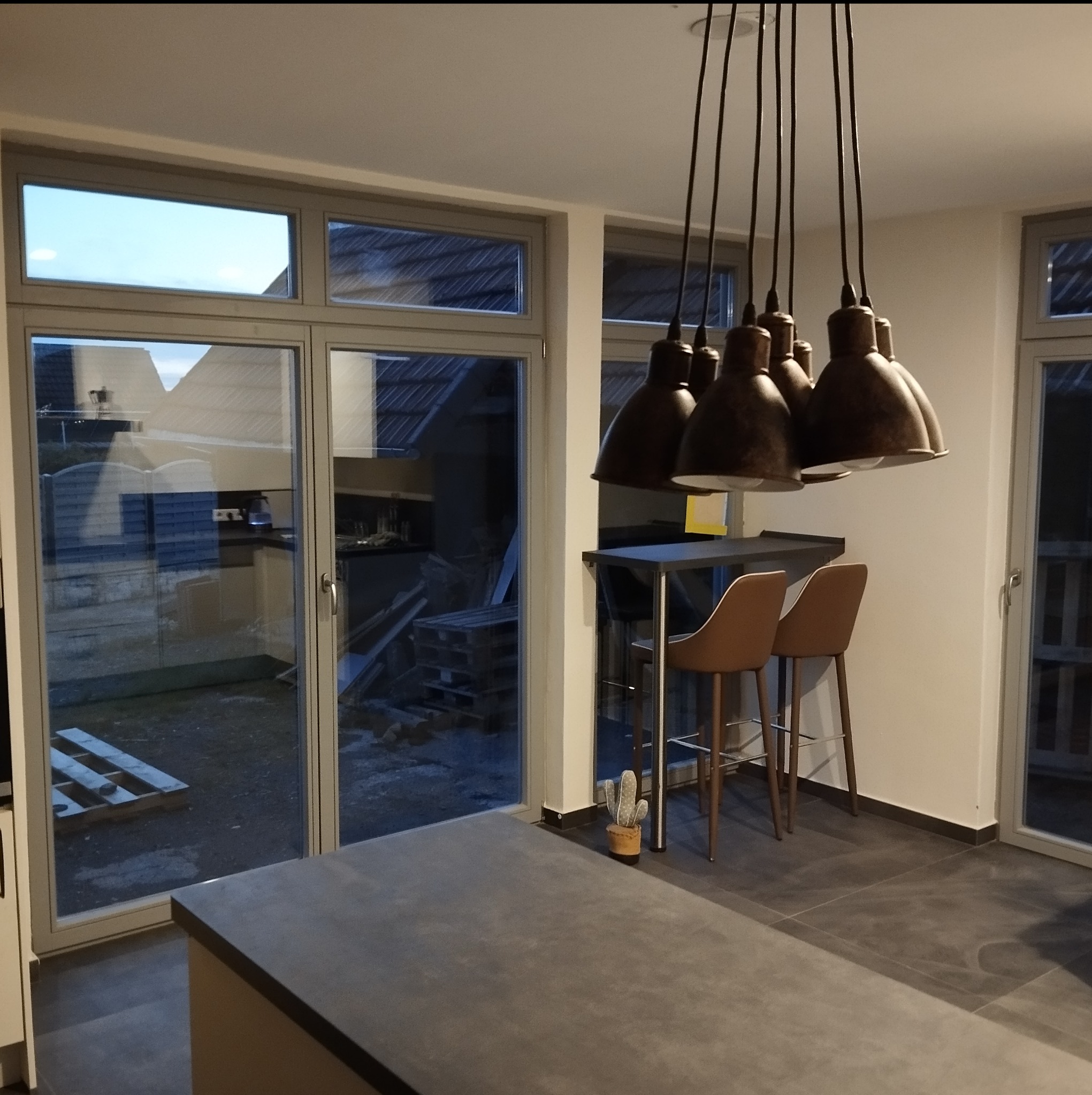 Nowoczesna kuchnia z szarym blatem, wysokimi krzesłami barowymi w kolorze karmelowym i oryginalnym oświetleniem w postaci wiszących lamp w stylu industrialnym. Widok na zewnątrz przez duże okno...
