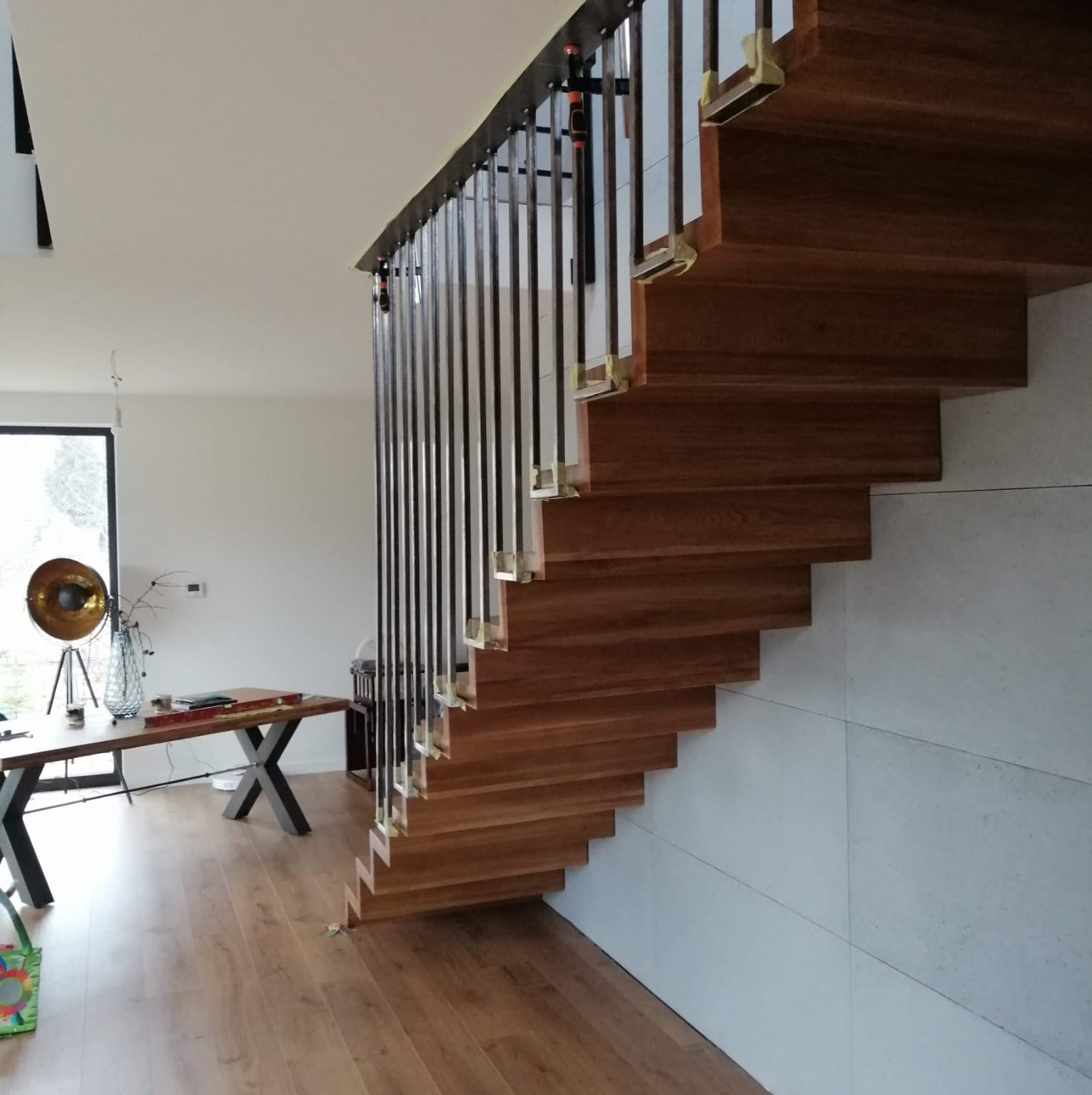 Nowoczesne, drewniane schody wiszące z czarną balustradą w jasnym wnętrzu z parkietem i minimalistycznym biurkiem.