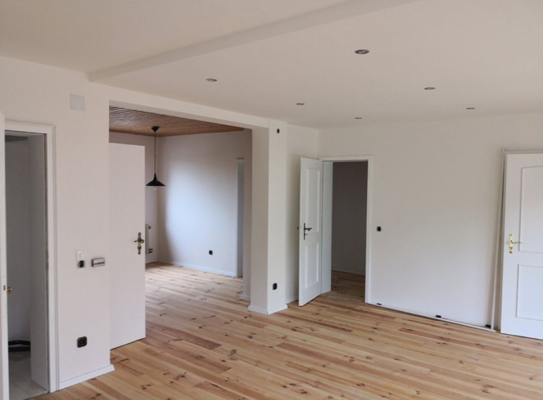 Jasne wnętrze po remoncie: świeżo pomalowane białe ściany, drewniana podłoga z widocznym usłojeniem, białe drzwi z czarnymi i złotymi klamkami, sufit z punktowym oświetleniem i drewnianym akcentem.