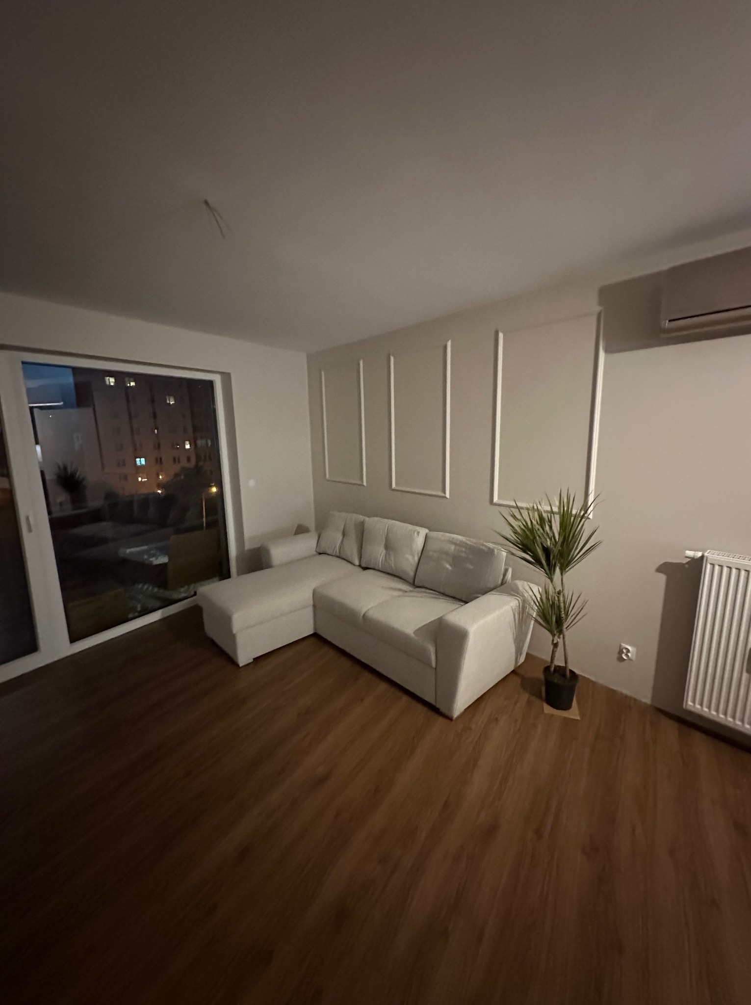 Jasnoszara narożna sofa z szezlongiem w nowocześnie urządzonym salonie z drewnianą podłogą i dekoracyjnymi listwami na ścianie, widok na oświetlone budynki za oknem.