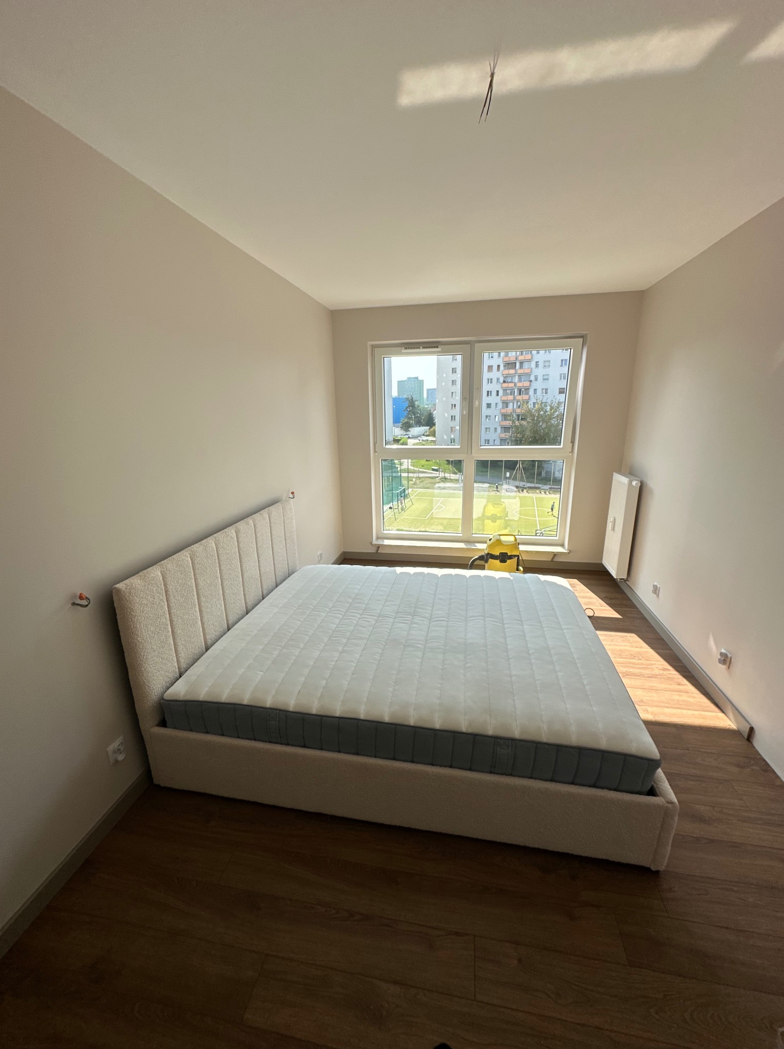 Nowo zmontowane łóżko tapicerowane z wełnianym zagłówkiem i materacem w minimalistycznej sypialni z parkietem, widok na boisko sportowe za oknem, widoczny odkurzacz na tle okna.