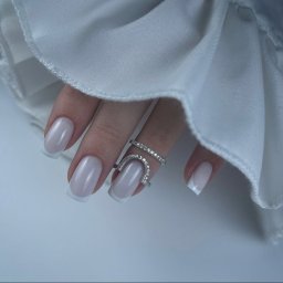 Nails_Beauty.LEGIONOWO - Dłoń z eleganckim, jasnym french manicure i dwoma srebrnymi pierścionkami z cyrkoniami na tle białej tkaniny.