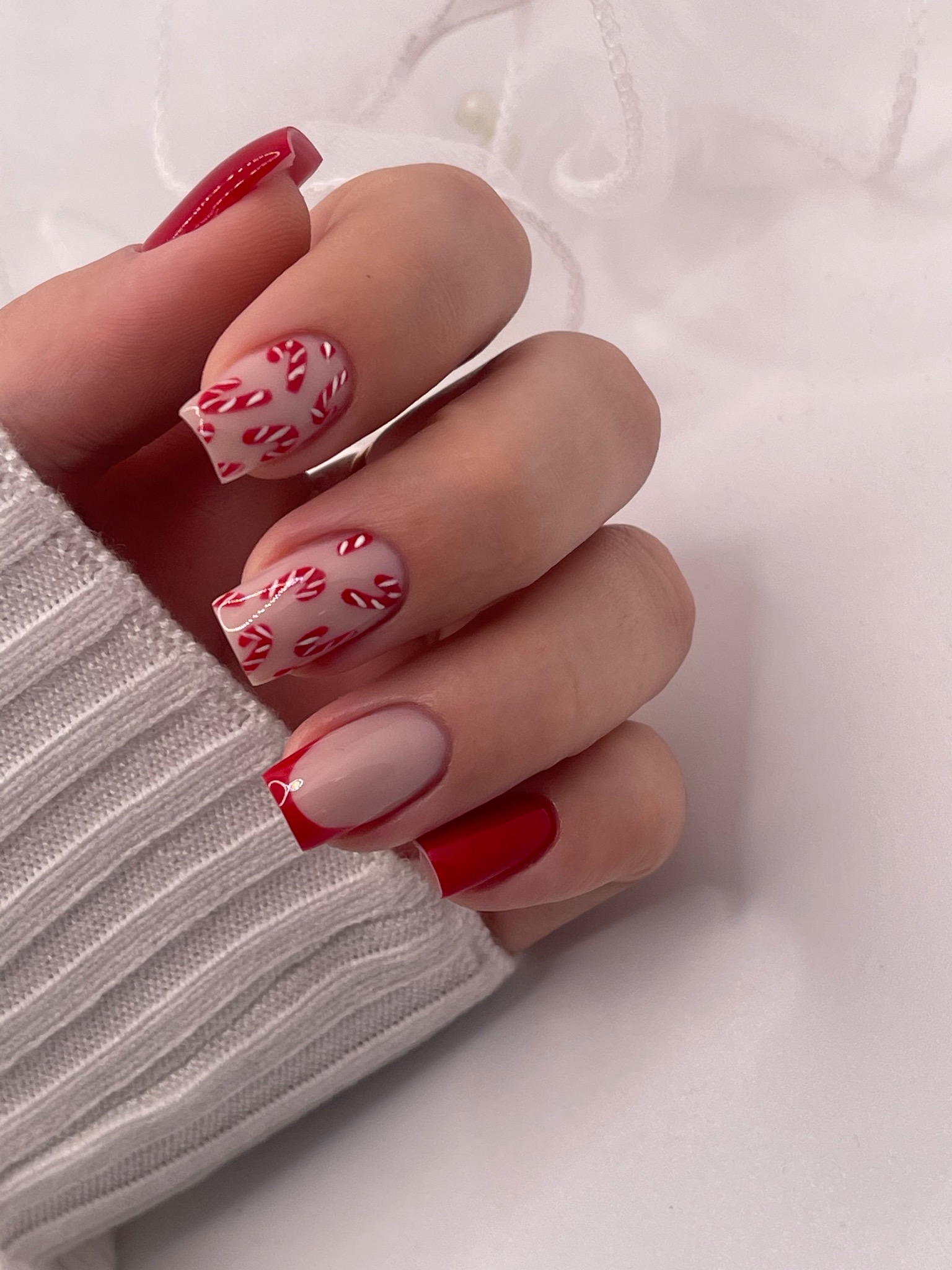 Dłoń z kwadratowymi paznokciami: jeden pomalowany na intensywny czerwony kolor, pozostałe z motywem czerwonych lasek cukrowych na transparentnym tle i czerwonym francuskim wykończeniem, na tle...