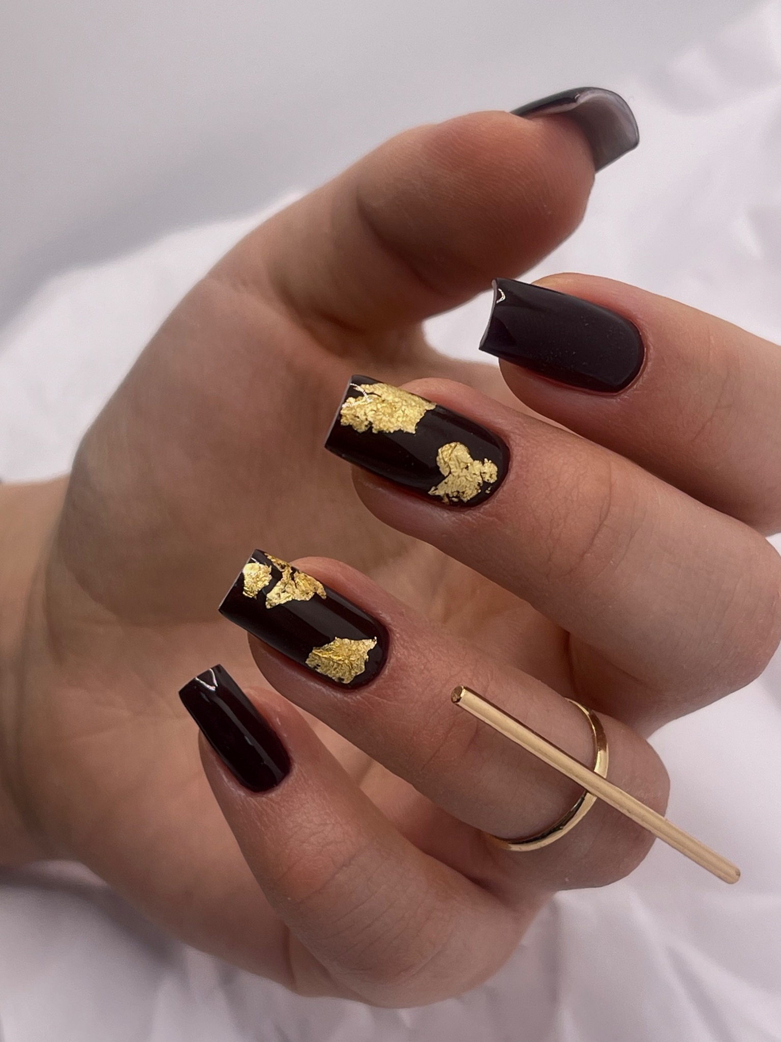 Dłoń z ciemnymi, kwadratowymi paznokciami ozdobionymi złotymi płatkami, uzupełniona złotym pierścionkiem z poprzeczką.