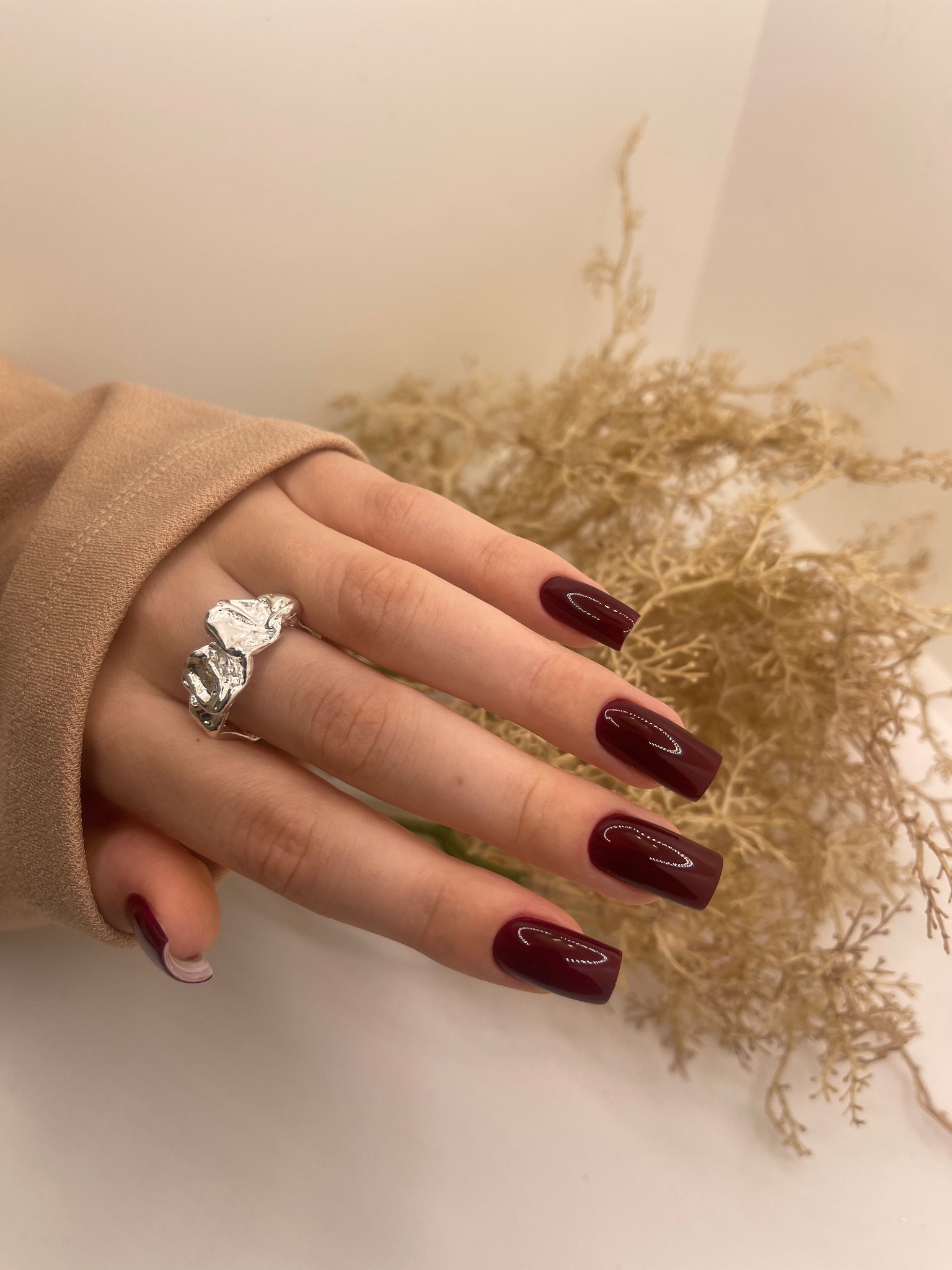 Dłoń z kwadratowymi paznokciami w kolorze głębokiej wiśni, ozdobiona dużym, srebrnym pierścieniem o organicznej formie, na tle delikatnych, suchych gałązek.