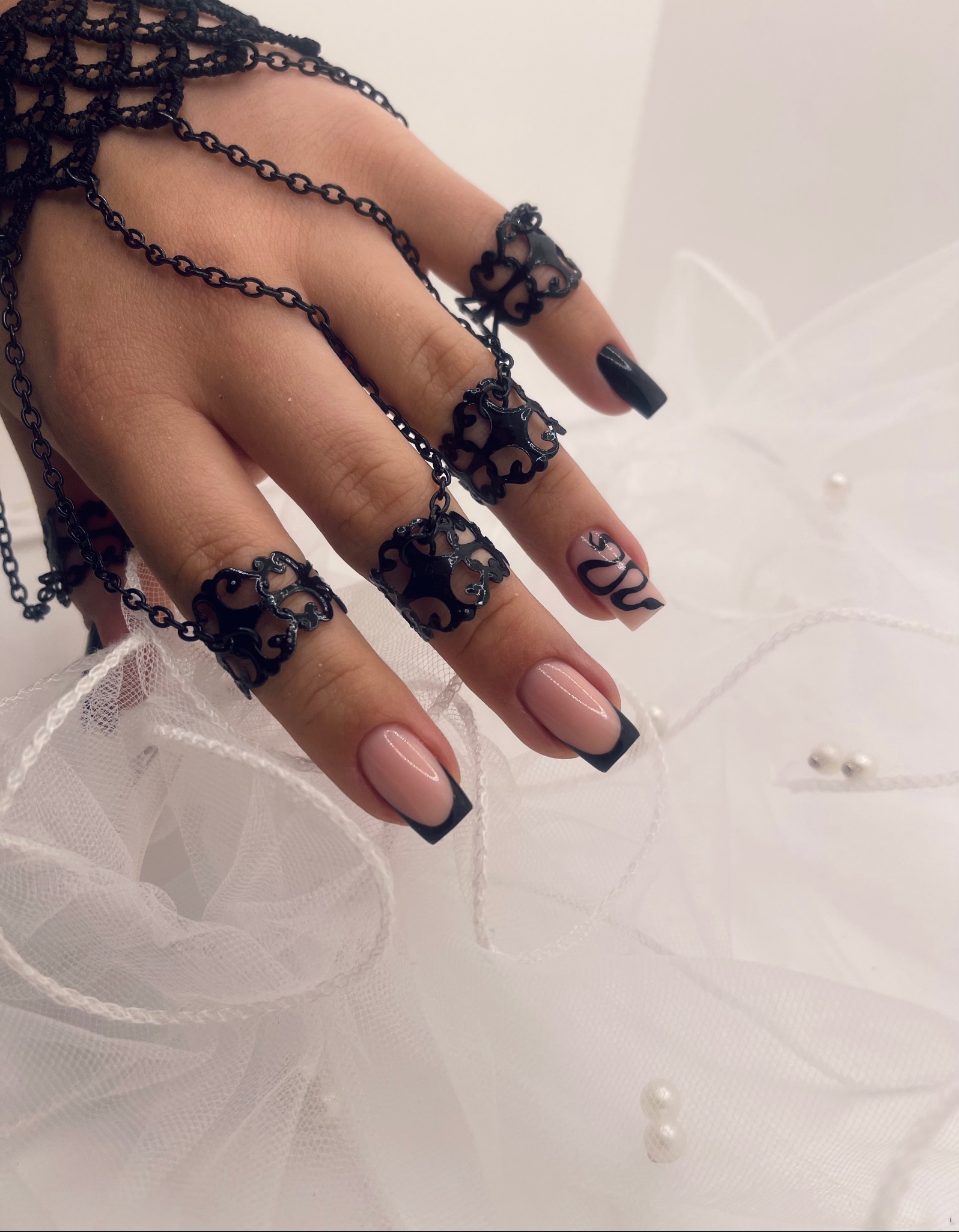 Dłoń z czarną, ażurową biżuterią na palcach, prezentująca french manicure z czarnymi końcówkami oraz zdobienie w postaci czarnego węża na jednym paznokciu, na tle białego tiulu z perłami.