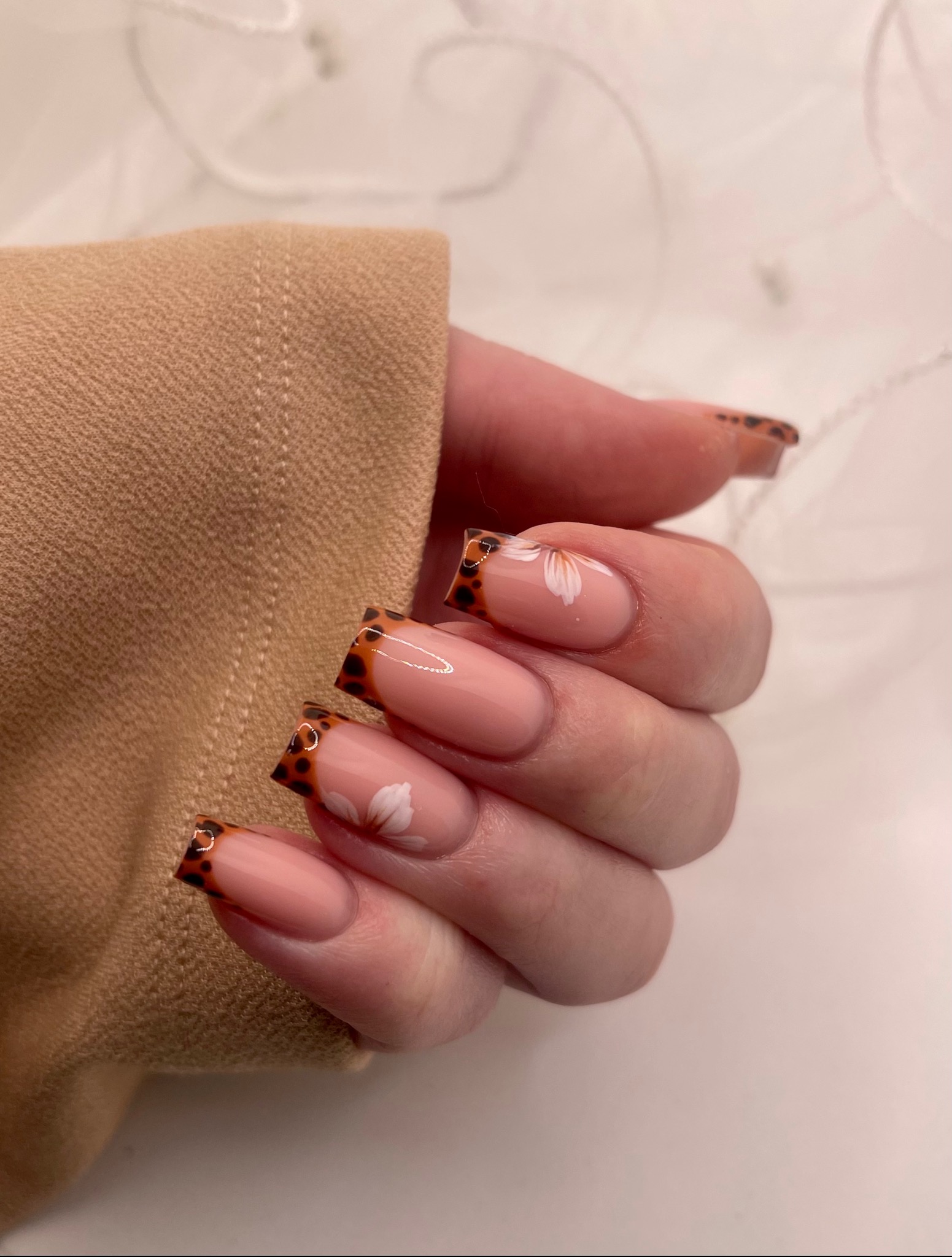 Dłoń z kwadratowymi paznokciami french w panterkę, zdobionymi białymi kwiatkami na beżowym tle bluzki i rozmytej firanki.