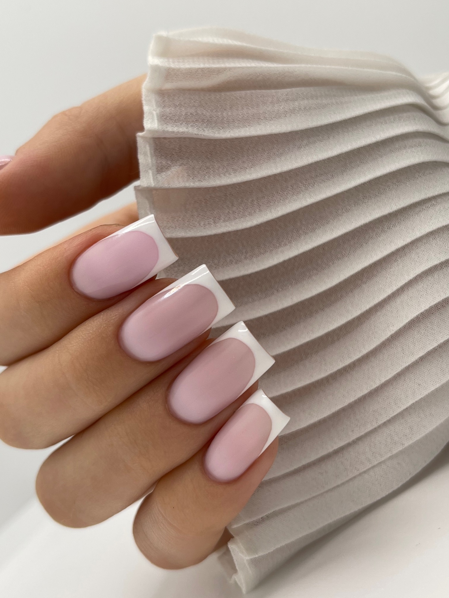 Dłoń z kwadratowymi paznokciami w delikatnym różu z białym french, na tle białej plisowanej tkaniny.