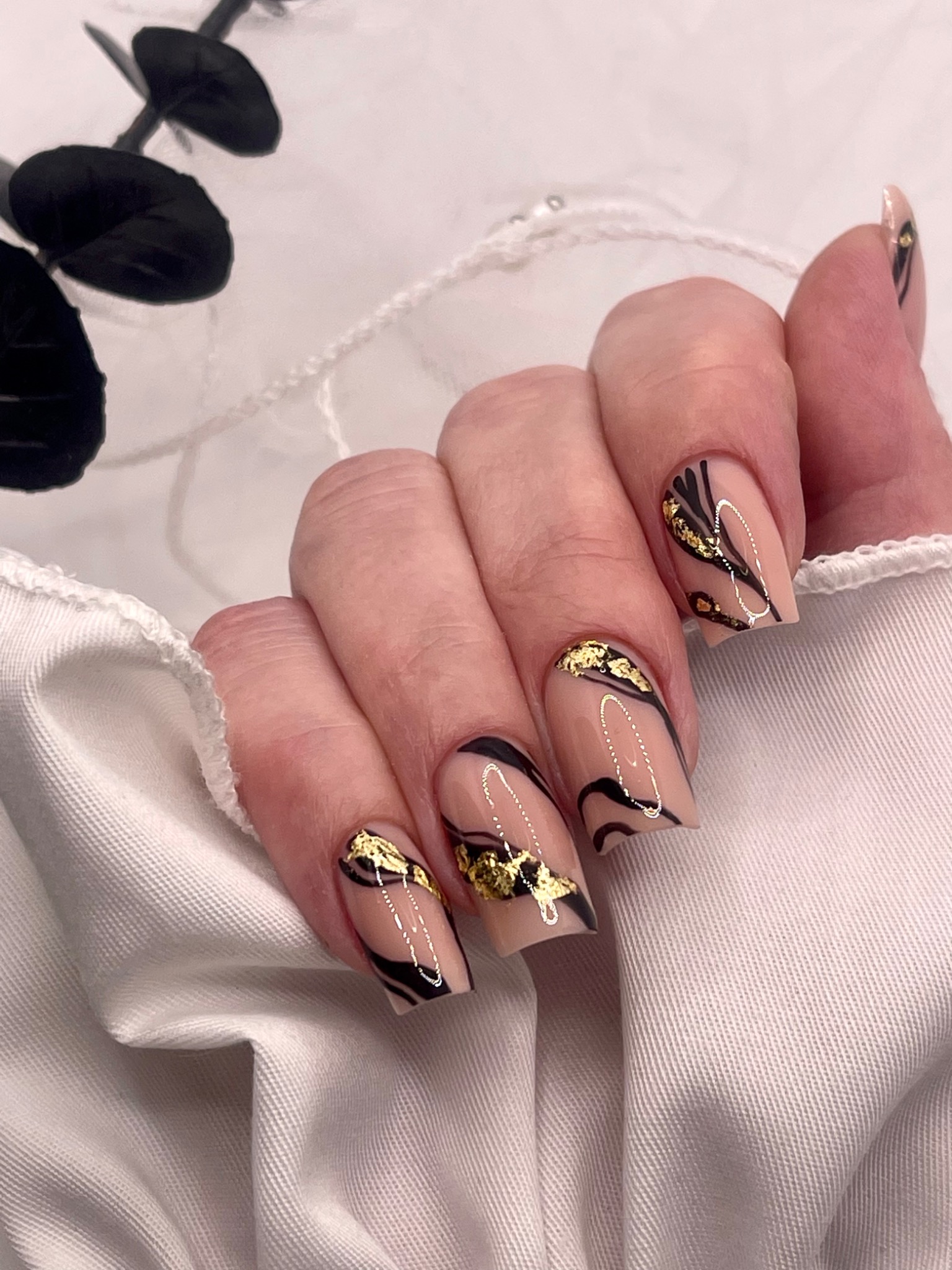 Dłoń z kwadratowymi paznokciami w odcieniach beżu, zdobionymi abstrakcyjnymi, czarnymi liniami, złotymi płatkami i srebrnymi akcentami na tle białej tkaniny i czarnych liści eukaliptusa.