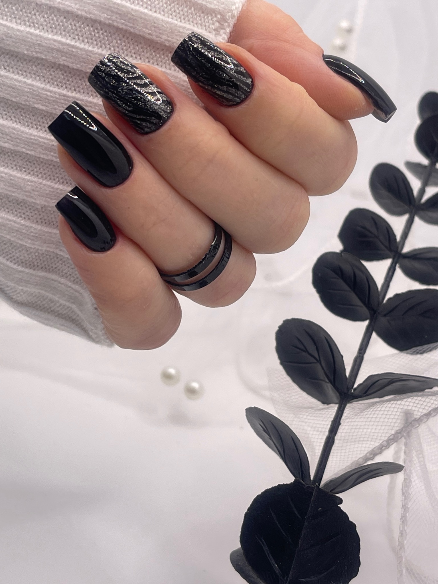 Dłoń z czarnymi, kwadratowymi paznokciami; dwa z nich zdobione srebrzystym wzorem, na tle białej tkaniny, czarnej dekoracyjnej gałązki i dwóch białych perełek.