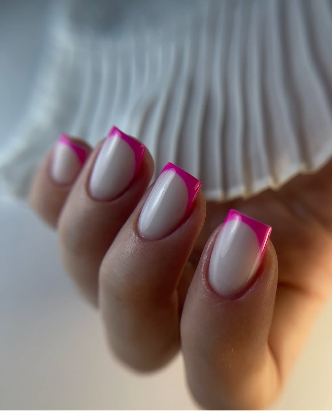 Dłoń z francuskim zdobieniem paznokci w kolorze białym i neonowym różowym, na tle białej, plisowanej tkaniny.