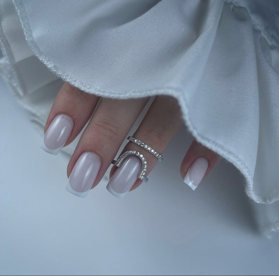 Dłoń z eleganckim, jasnym french manicure i dwoma srebrnymi pierścionkami z cyrkoniami na tle białej tkaniny.