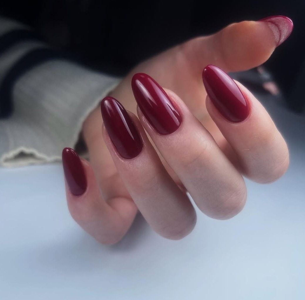 Dłoń z pomalowanymi na ciemny burgund, błyszczącymi paznokciami w kształcie migdała na jasnym tle.