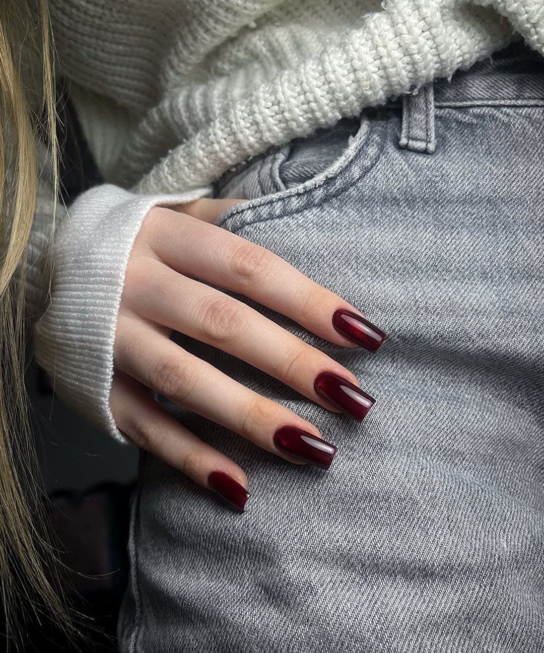 Dłoń z ciemnoczerwonymi, kwadratowymi paznokciami na tle szarych jeansów i białego swetra.