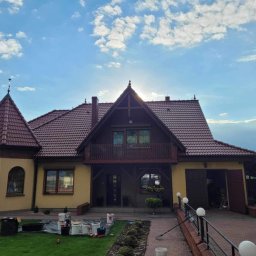 Malowanie dachów Szymon Przychodzki - Żółty dom jednorodzinny z brązowym dachem z dachówki, widoczne puszki z farbą i akcesoria malarskie na trawniku przed domem, sugerujące prace malarskie w toku.