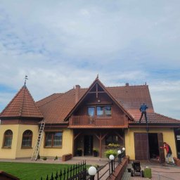 Malowanie dachów Szymon Przychodzki - Dwóch mężczyzn czyści dach dużego, dwupiętrowego domu z czerwonej dachówki za pomocą myjki ciśnieniowej; jeden stoi na dachu, drugi obsługuje urządzenie na ziemi.