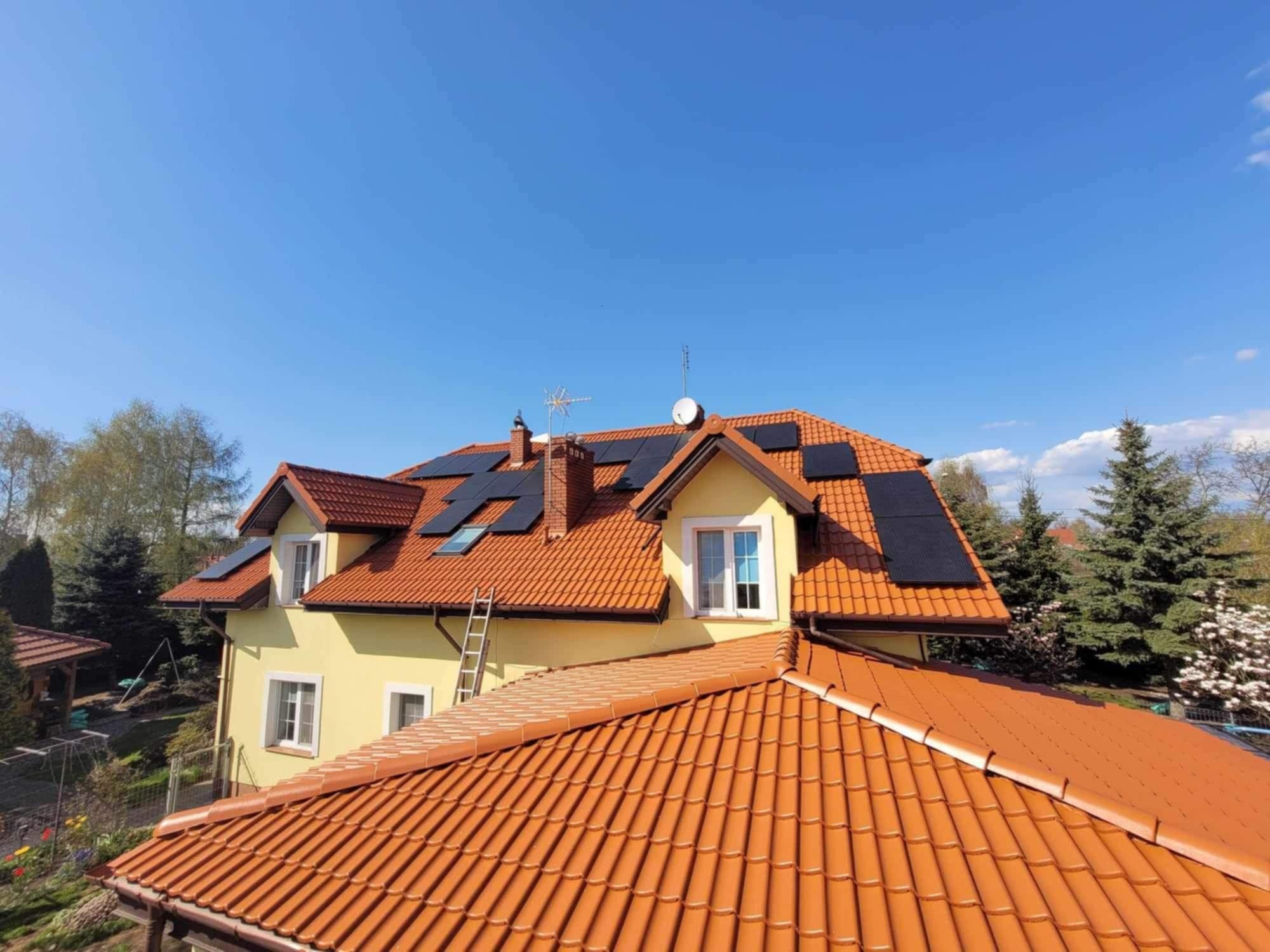 Żółty dom z pomarańczową dachówką i panelami słonecznymi na dachu, drabina oparta o ścianę, widok z góry w słoneczny dzień.