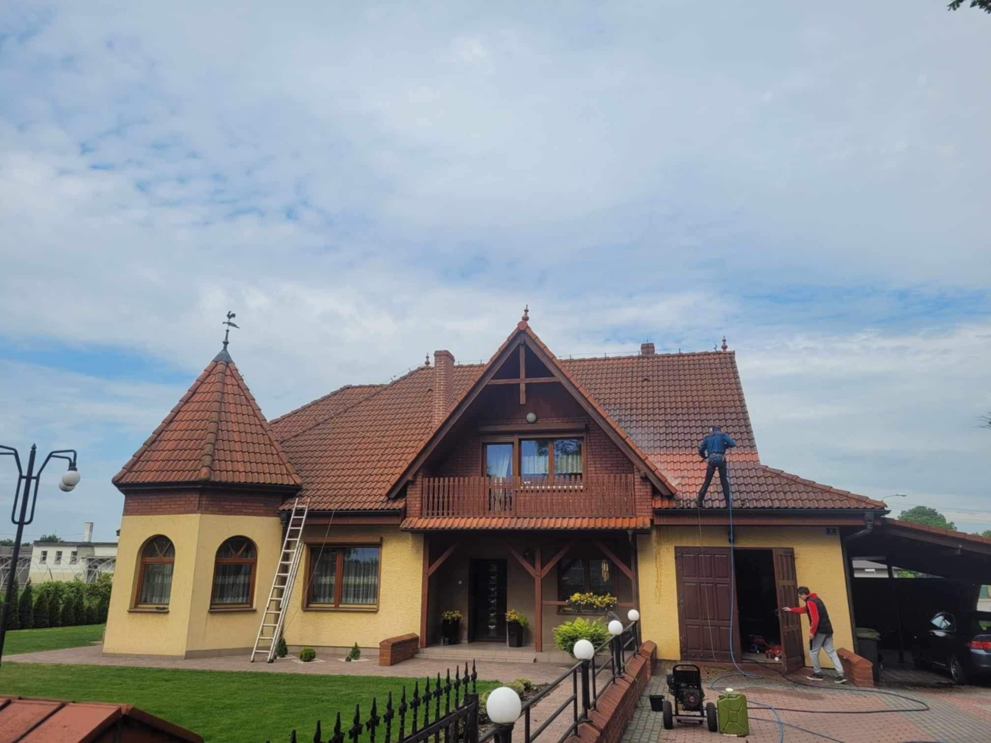 Dwóch mężczyzn czyści dach dużego, dwupiętrowego domu z czerwonej dachówki za pomocą myjki ciśnieniowej; jeden stoi na dachu, drugi obsługuje urządzenie na ziemi.