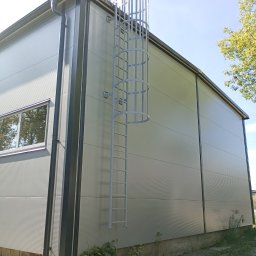 Damian Kacała - Hala przemysłowa obłożona płytami warstwowymi z zamontowaną zewnętrzną metalową drabinką z zabezpieczeniem koszowym, widok z dołu na fragment budynku.