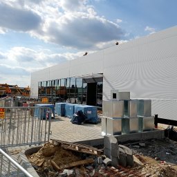 Damian Kacała - Budynek hali produkcyjnej w trakcie montażu płyt warstwowych, widoczne zabezpieczenia budowy, niebieskie paczki materiałów i stos metalowych kanałów wentylacyjnych.