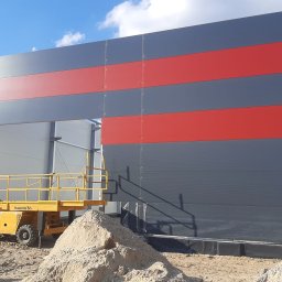 Damian Kacała - Montaż ściany hali z płyt warstwowych w kolorach szarym i czerwonym, widoczny podnośnik koszowy Haulotte H12S XL i pryzma piasku.