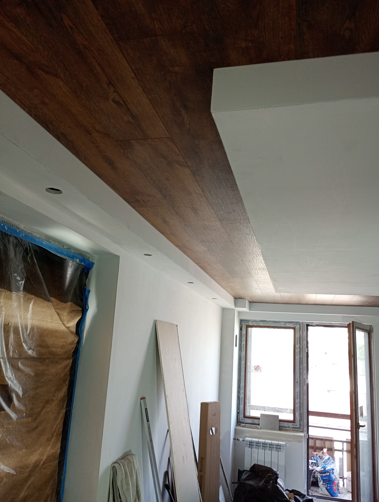 Wykończenie wnętrza: Laminowane panele na suficie, kontrastujące z białymi ścianami i wystającymi elementami sufitu. Widoczne okno z otwartym balkonem, zabezpieczone folią ściany oraz materiały...