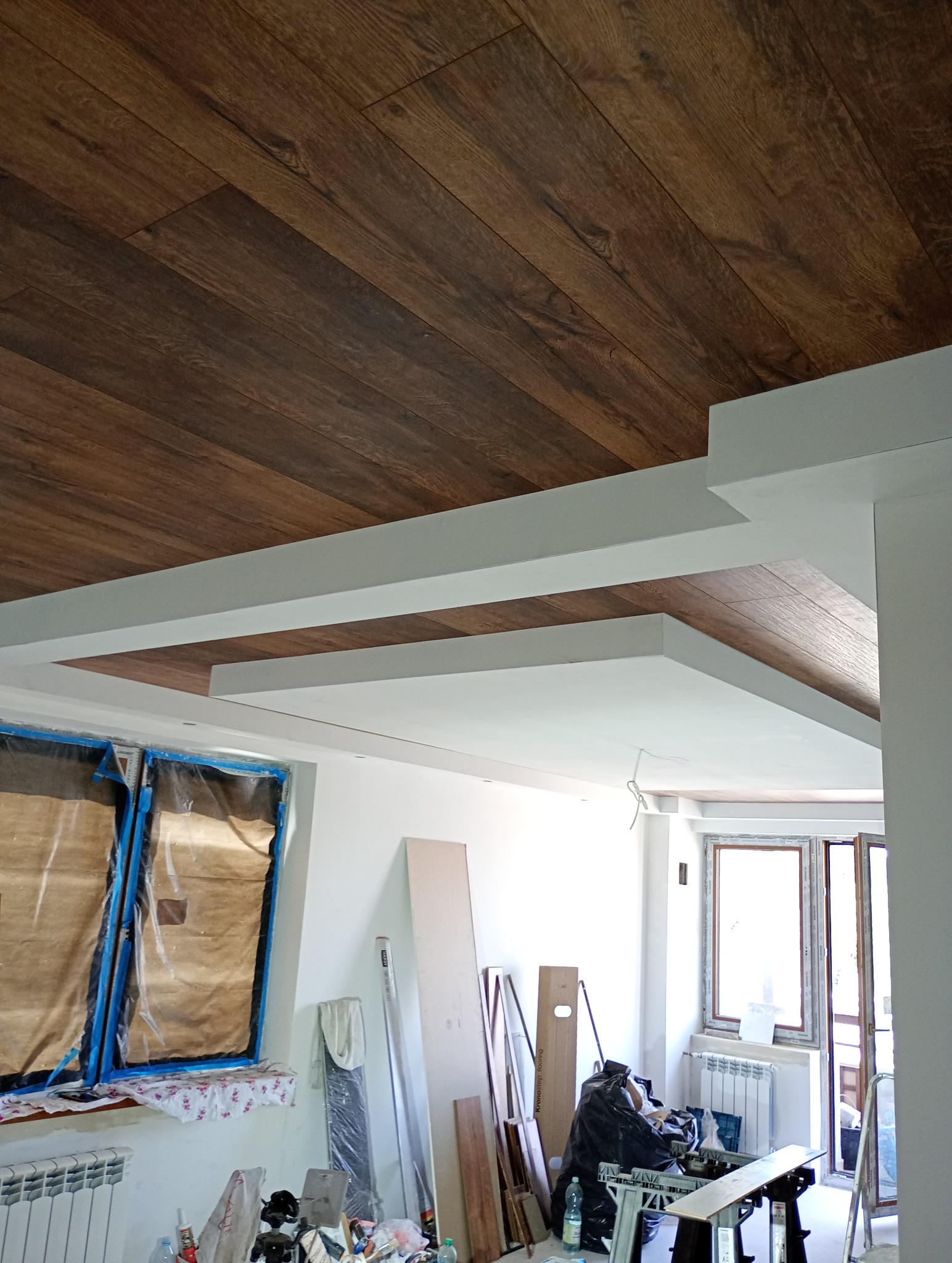 Drewnopodobna okładzina sufitowa w trakcie montażu, widoczne białe elementy konstrukcyjne i zabezpieczone okna, wnętrze w stanie surowym z narzędziami i materiałami budowlanymi.