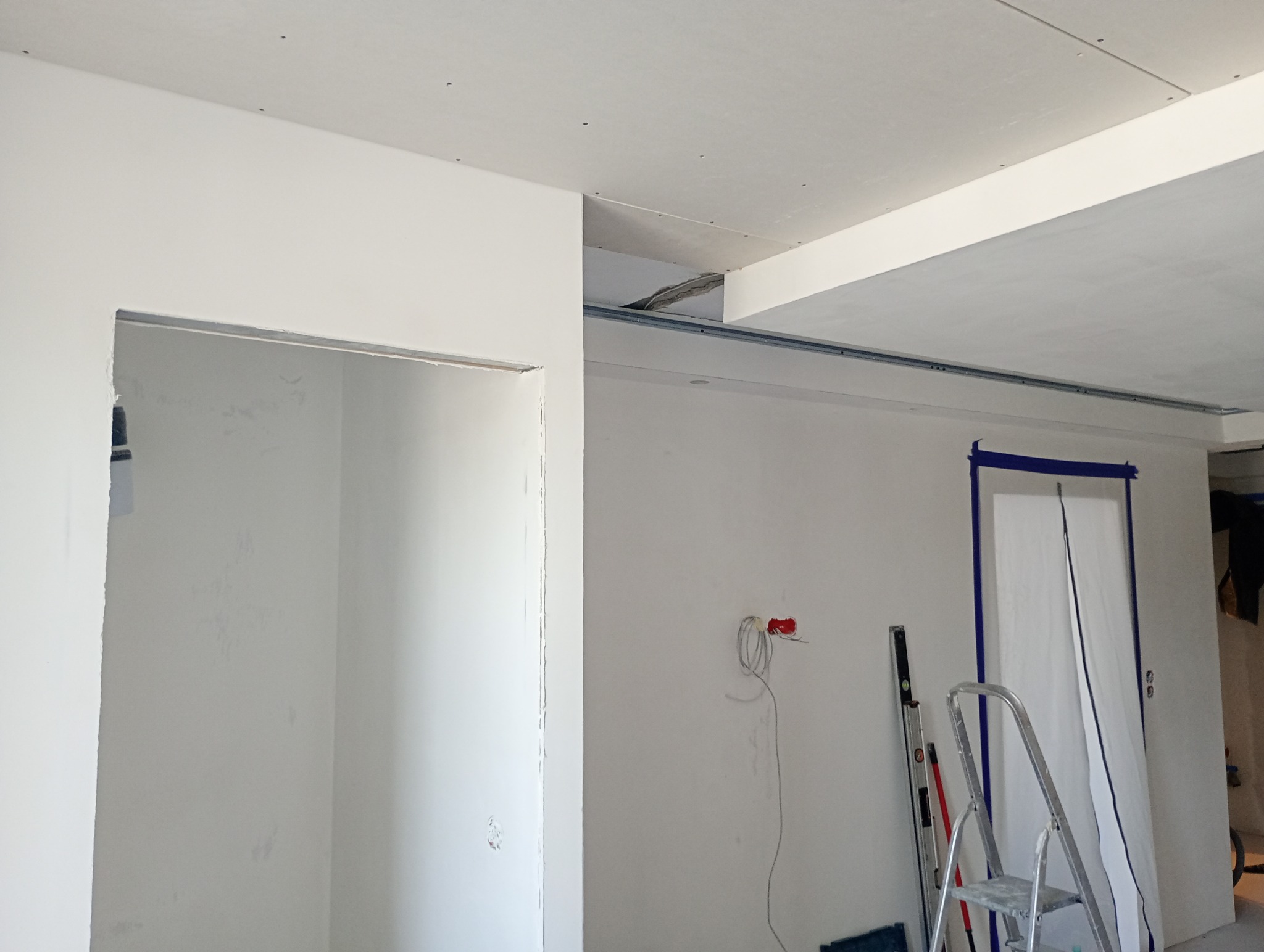Wnętrze w trakcie prac wykończeniowych: surowe ściany pomalowane na biało, widoczne przewody elektryczne, poziomica oparta o ścianę, drabina i zabezpieczone folią drzwi z niebieską taśmą malarską...