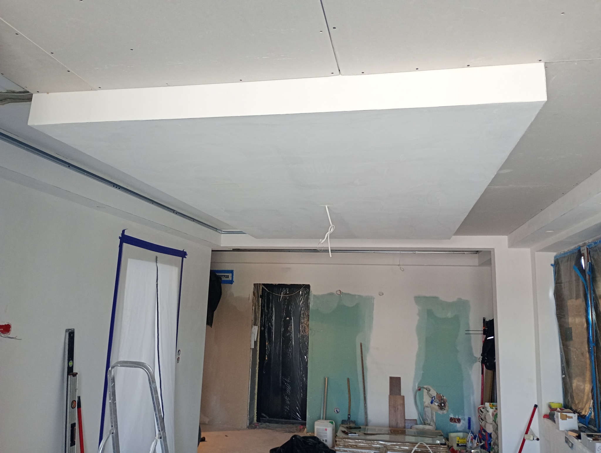 Sufit podwieszany z płyt gipsowo-kartonowych w trakcie montażu, widoczne profile i instalacja elektryczna, zabezpieczone drzwi folią malarską, ściany w trakcie szpachlowania, drabina i poziomica...
