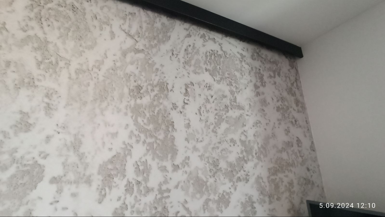 Ściana z dekoracyjną, nieregularną fakturą w odcieniach szarości i bieli, wykończona czarną listwą przysufitową.