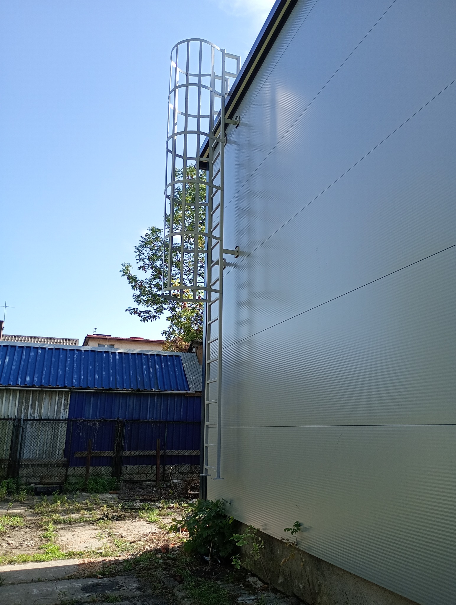 Zewnętrzna ściana hali przemysłowej z blachy trapezowej w kolorze jasnoszarym, wyposażona w metalową drabinę z osłoną, prowadzącą na dach; widoczne są fragmenty sąsiednich budynków i zarośli.