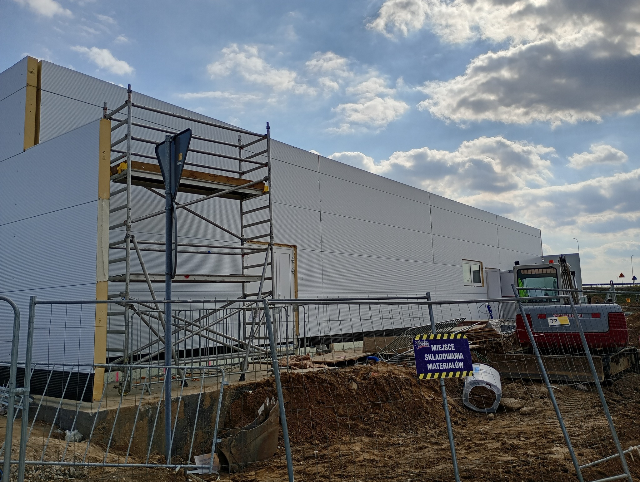 Nowo wznoszona hala z płyt warstwowych, widoczny rusztowanie przy elewacji, teren budowy zabezpieczony metalowym ogrodzeniem, w tle błękitne niebo z chmurami.