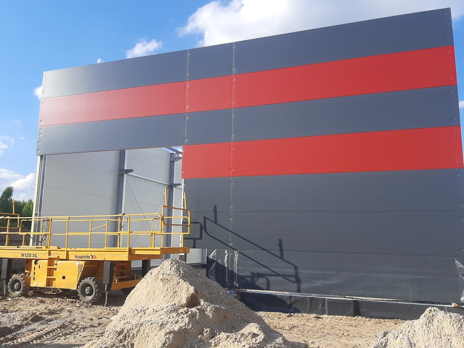 Montaż ściany hali z płyt warstwowych w kolorach szarym i czerwonym, widoczny podnośnik koszowy Haulotte H12S XL i pryzma piasku.