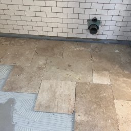 Filip W - Układanie beżowych, kwadratowych płytek podłogowych w łazience, widoczny fragment ściany z białymi, prostokątnymi płytkami, szare fugi, plastikowe krzyżyki dystansowe, świeży klej do płytek