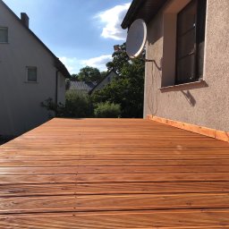 Filip W - Nowa, drewniana podłoga tarasu z ryflowanych desek w ciepłym odcieniu brązu, widok z niskiej perspektywy, zarys budynków w tle i antena satelitarna na elewacji.