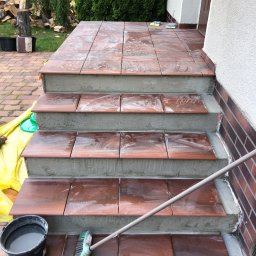 Filip W - Układanie brązowych płytek na schodach wejściowych, widoczne wiadro z wodą i szczotka do czyszczenia, częściowo zabezpieczone folią malarską.