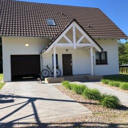 Filip W - Biały dom jednorodzinny z brązowym dachem, podjazd z szarej kostki brukowej, rower oparty o wejście, mały pies na podjeździe.