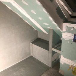 Filip W - Łazienka na poddaszu w trakcie prac wykończeniowych: ściany pokryte szarą warstwą, sufit z płyt gipsowo-kartonowych, wnęka z półkami w stanie surowym, widoczne elementy instalacji hydraulicznej.