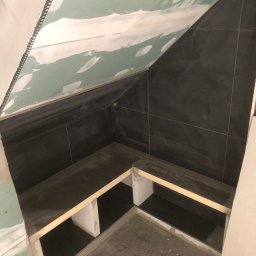 Filip W - Niedokończona łazienka na poddaszu: czarne płytki na ścianach, surowy gips na skosach, konstrukcja półek z płytek i drewnianych belek, surowa podłoga.