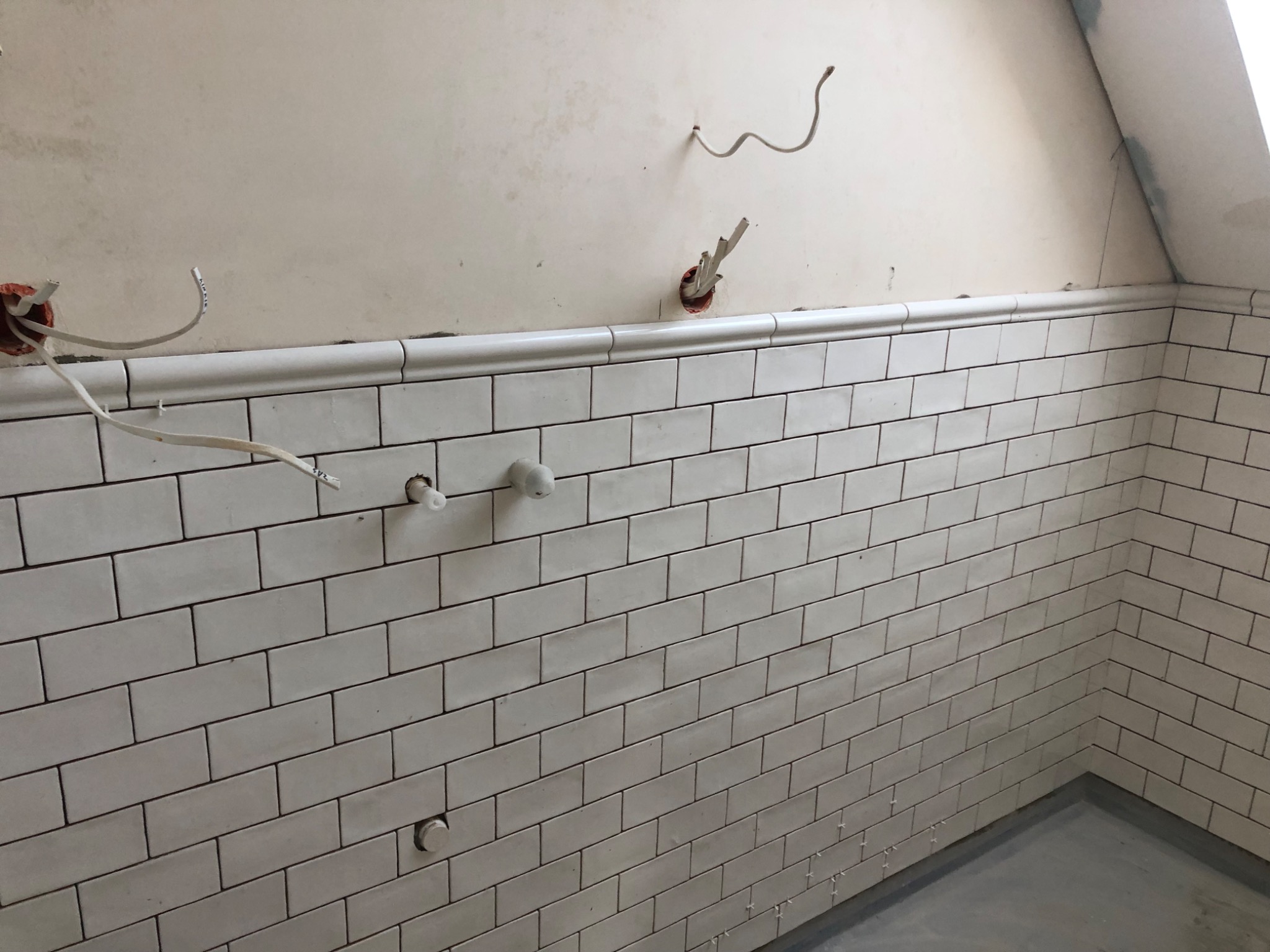 Ściana łazienki w trakcie remontu z ułożonymi białymi, prostokątnymi płytkami i wystającymi przewodami elektrycznymi ze ściany.