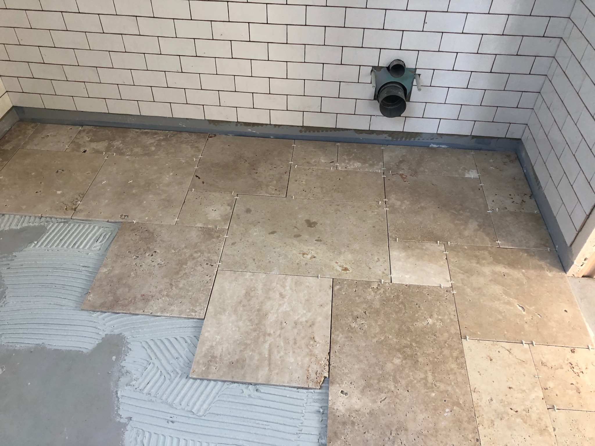 Układanie beżowych, kwadratowych płytek podłogowych w łazience, widoczny fragment ściany z białymi, prostokątnymi płytkami, szare fugi, plastikowe krzyżyki dystansowe, świeży klej do płytek