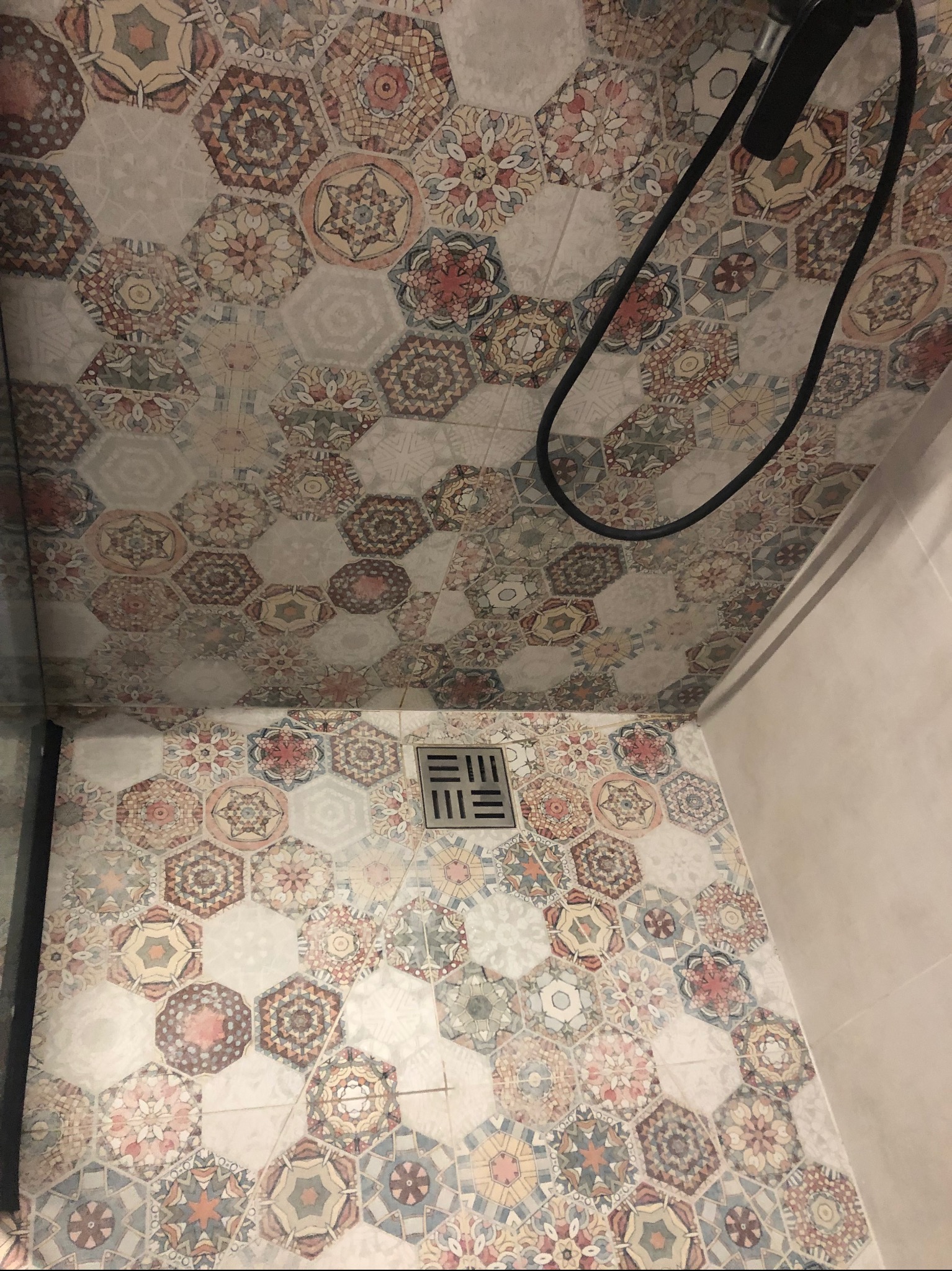 Wykończona łazienka z dekoracyjnymi, heksagonalnymi płytkami na podłodze i suficie; czarny wąż prysznicowy zwisa z sufitu; odpływ liniowy w podłodze.