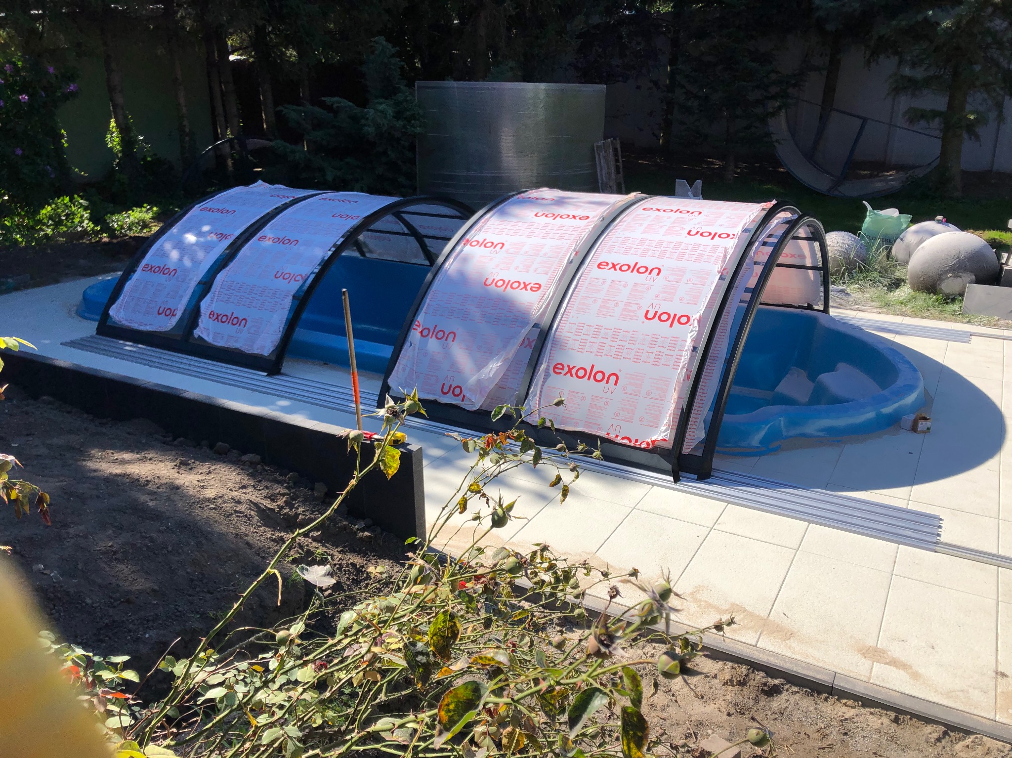 Niebieski basen ogrodowy z częściowo zamontowanym zadaszeniem z poliwęglanu Exolon, widoczne elementy montażowe i przygotowany teren wokół.