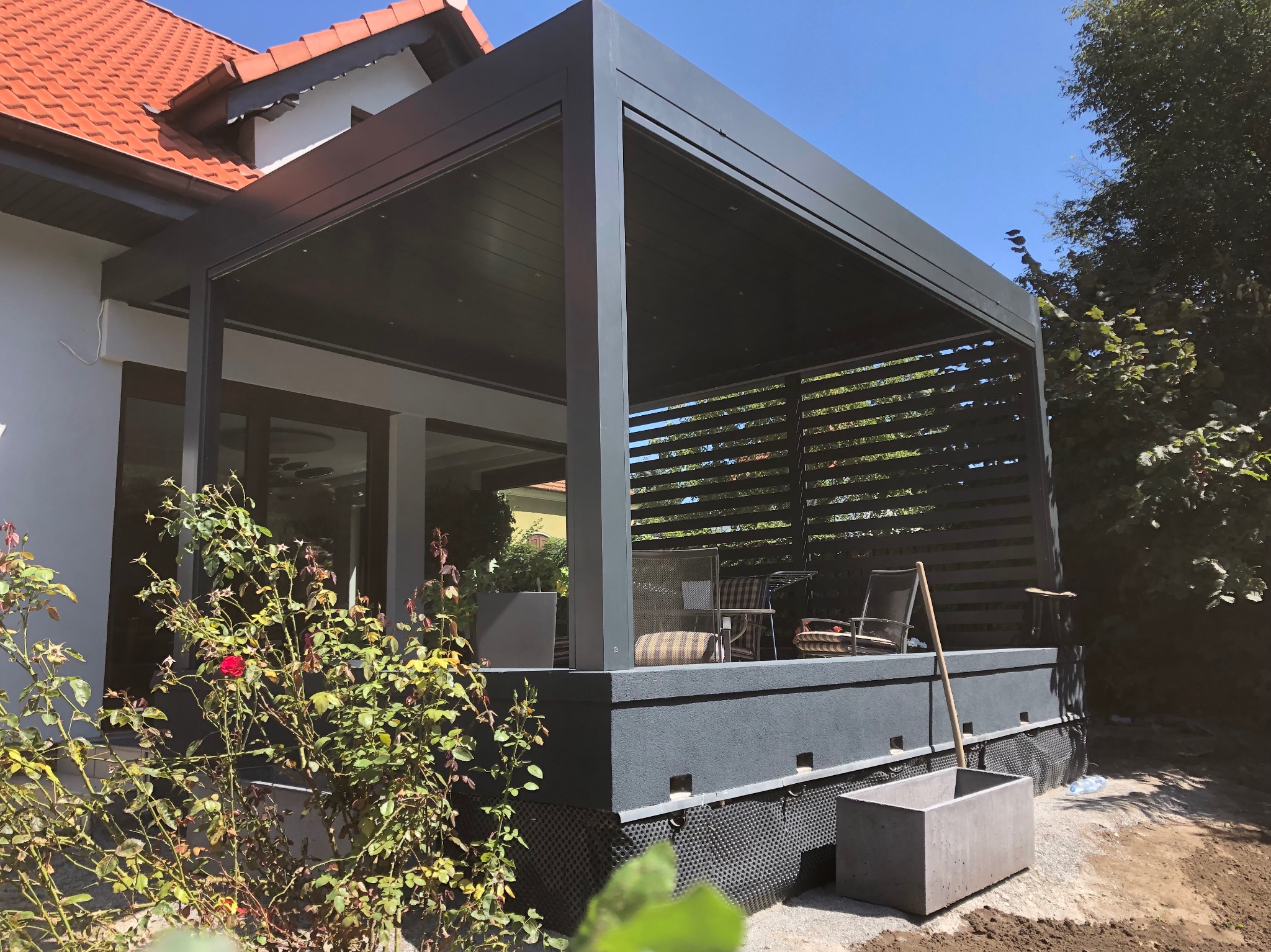 Nowoczesna, szara pergola z zadaszeniem lamelowym, widok z zewnątrz na taras z meblami ogrodowymi, w tle dom z czerwoną dachówką i błękitne niebo.