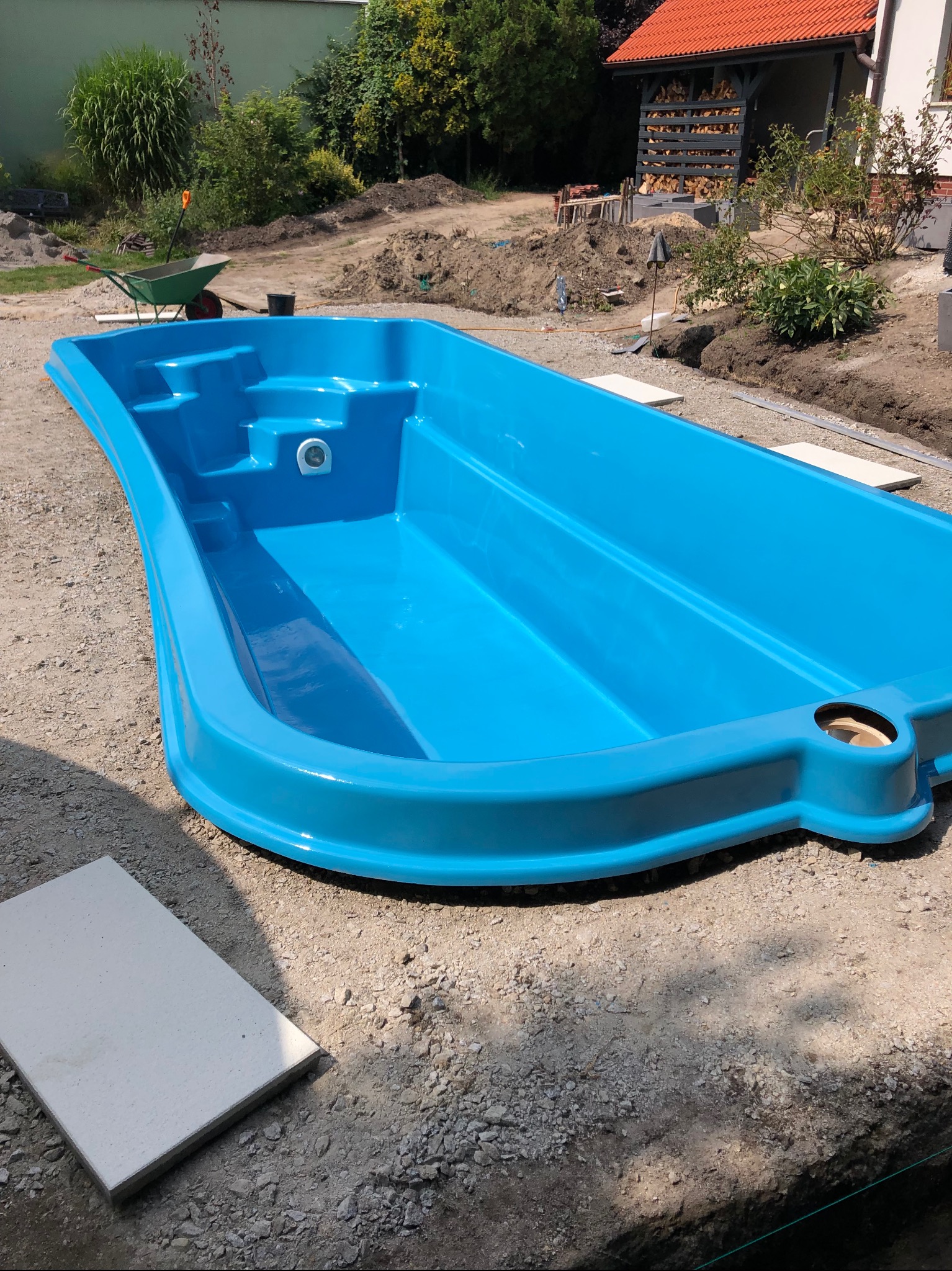Niebieski, gotowy basen z tworzywa sztucznego umieszczony w wykopie na działce, przygotowany do montażu, widoczne narzędzia i materiały budowlane w tle.