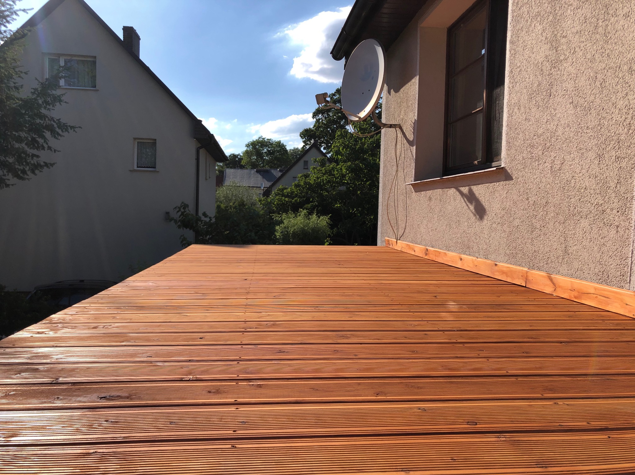 Nowa, drewniana podłoga tarasu z ryflowanych desek w ciepłym odcieniu brązu, widok z niskiej perspektywy, zarys budynków w tle i antena satelitarna na elewacji.