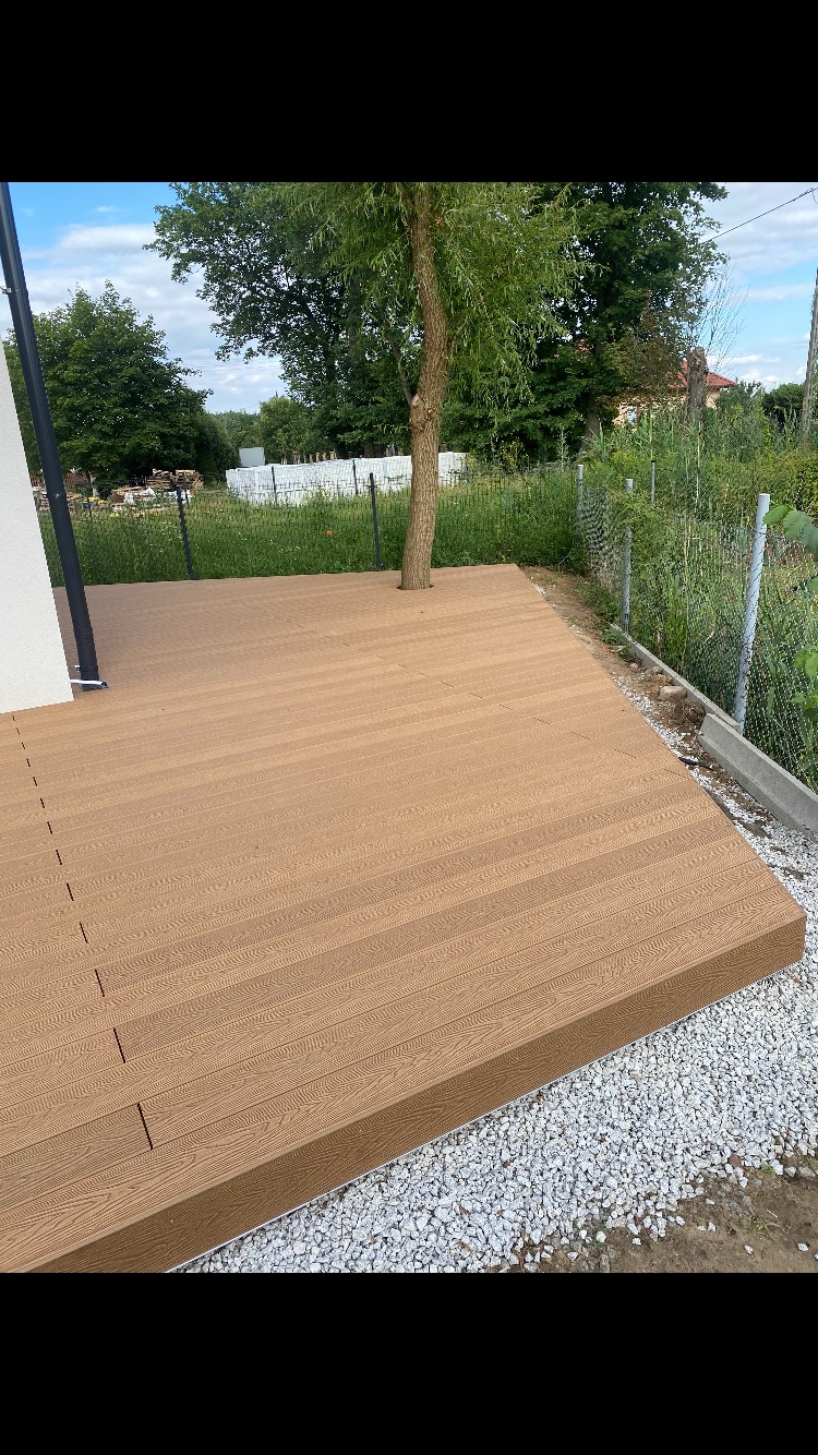Nowoczesny taras z desek kompozytowych w kolorze jasnego drewna, z wkomponowanym drzewem, otoczony żwirem i zielenią.