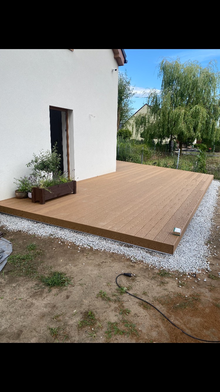 Nowo wybudowany taras z desek kompozytowych w kolorze brązowym, otoczony drobnym żwirem, przylegający do białego budynku z otwartymi drzwiami i donicą z roślinami.