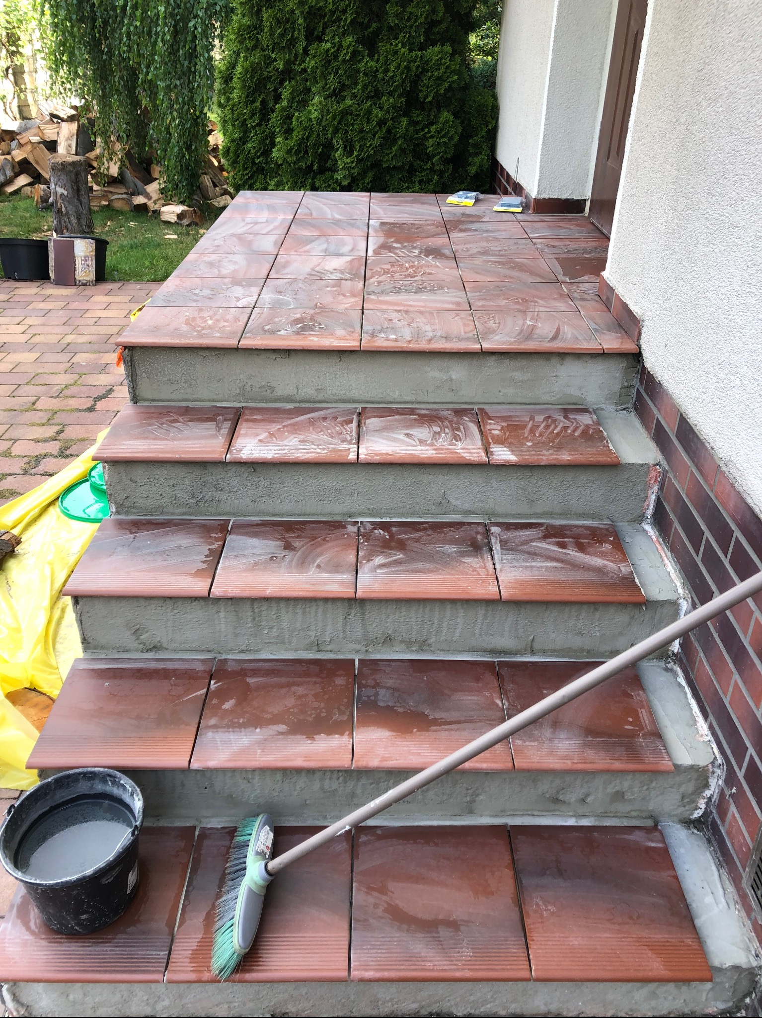 Układanie brązowych płytek na schodach wejściowych, widoczne wiadro z wodą i szczotka do czyszczenia, częściowo zabezpieczone folią malarską.