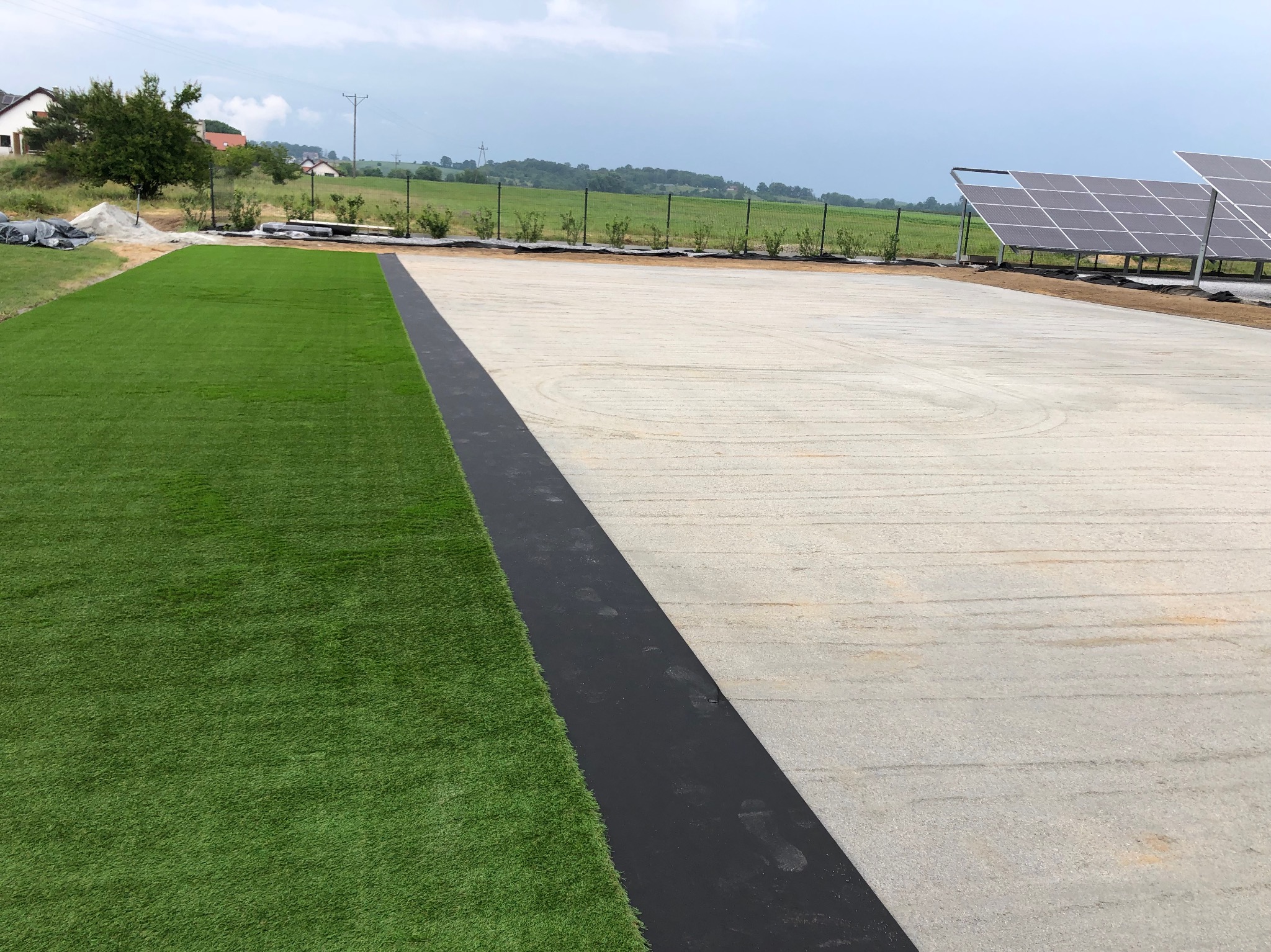 Widok z góry na przygotowane podłoże z ułożoną sztuczną trawą po lewej stronie i betonową wylewką po prawej, oddzielone czarnym pasem, w tle panele słoneczne i zieleń.